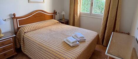 A hotel room with a large bed, a nightstand, a lamp, an air conditioning unit, a window with curtains, and a painting on the wall.