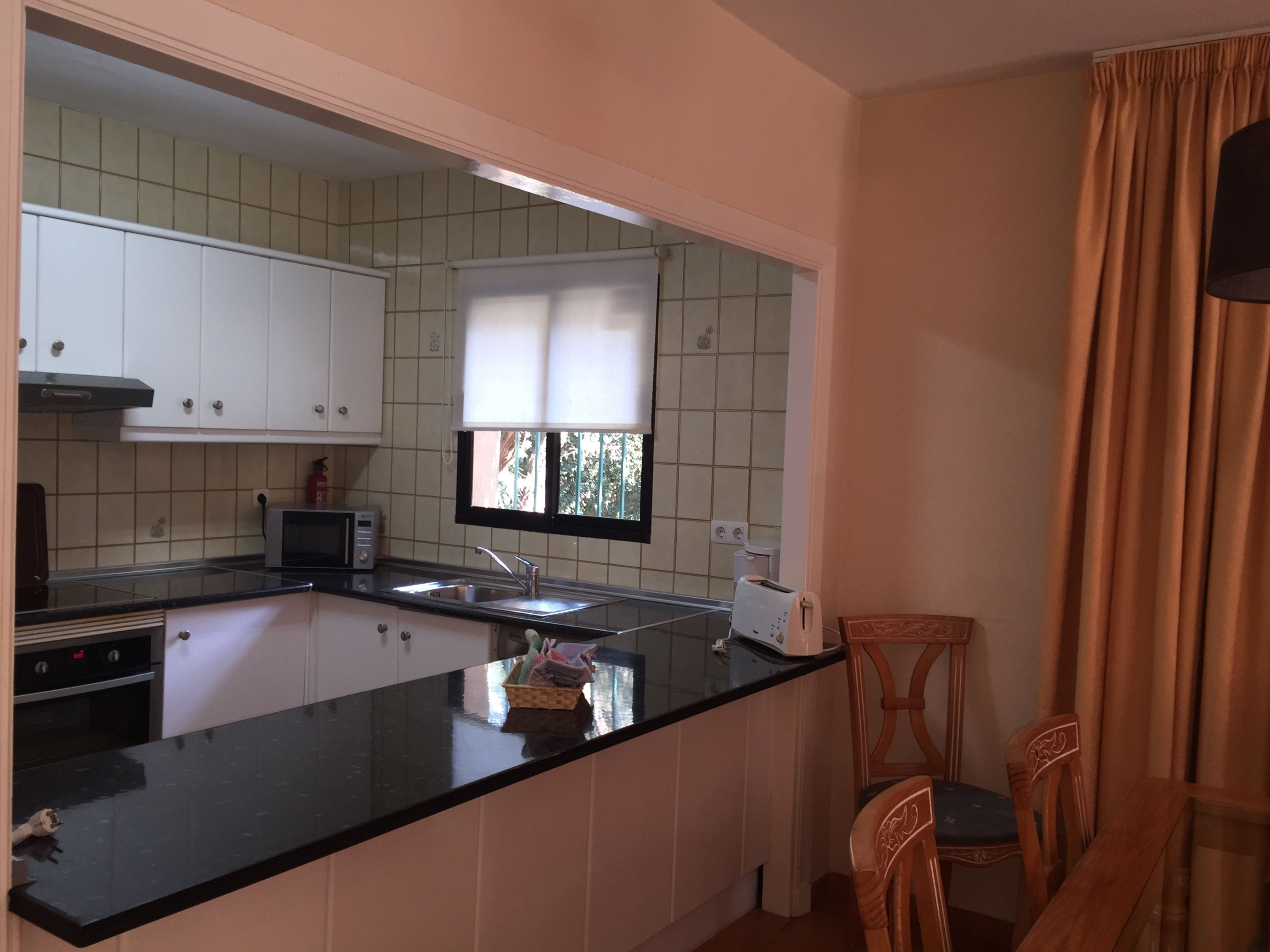 A kitchen with white cabinets, a black countertop, a window with blinds, and a dining area with wooden chairs.