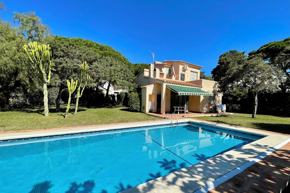 A swimming pool with a tiled deck, a covered patio with outdoor furniture, and a two-story building with a tiled roof and a green awning.