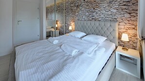 A bedroom with a stone wall, a tufted headboard, a white bed with rolled towels, and bedside lamps.