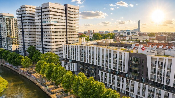 A high-rise building with multiple balconies, a river in the foreground, and a clear sky.