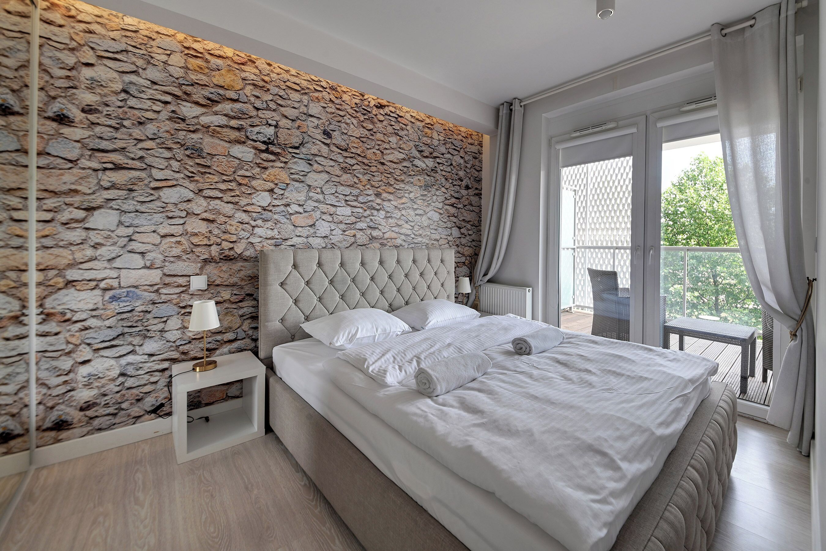 Une chambre avec un mur en pierre, une tête de lit capitonnée, un lit avec du linge de lit blanc, une table de chevet avec une lampe et une vue sur l’extérieur grâce à une porte coulissante.
