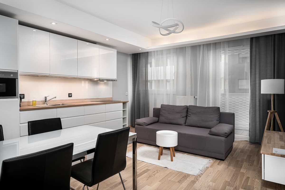 A modern living room with a grey sofa, a small white ottoman, and a dining area with a white table and black chairs.