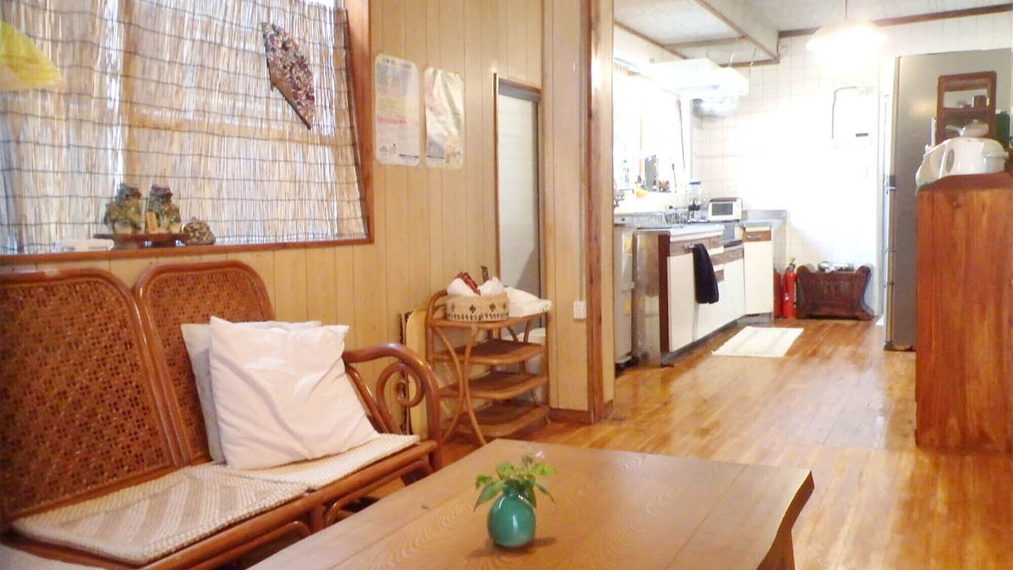 A room with a wooden sofa, a dining table with a vase, and a kitchen area with a microwave.