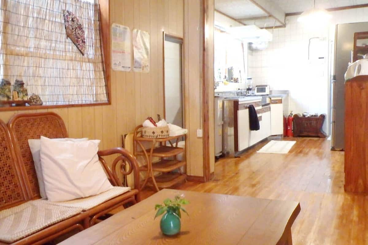 A room with a wooden sofa, a dining table with a vase, and a kitchen area with a microwave.