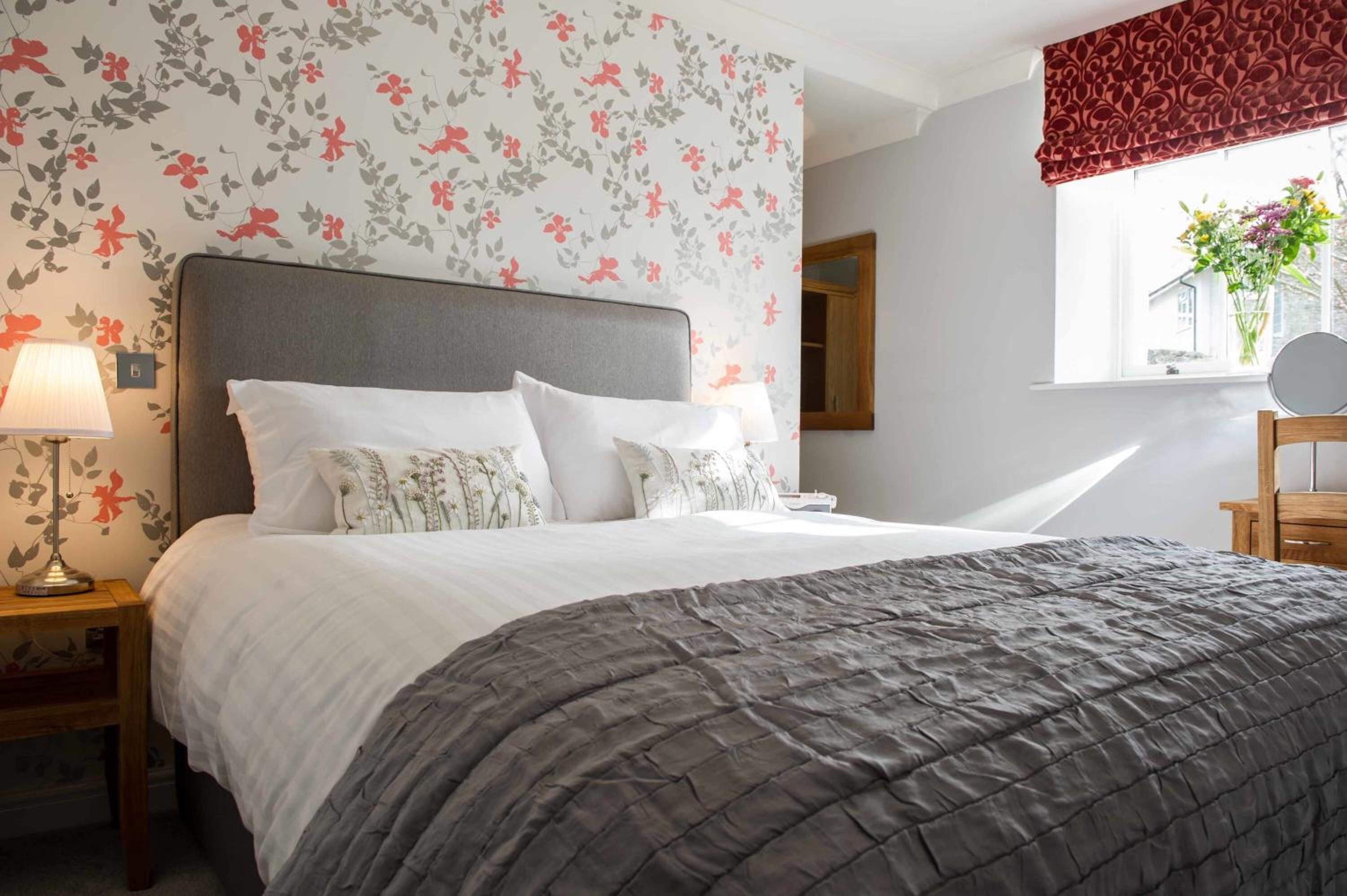 A neatly made bed with a grey quilt, a floral wallpaper pattern, and a window with a red valance.