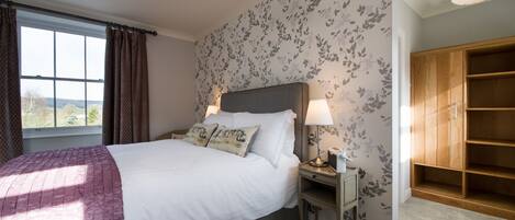 A bedroom with a large bed, a nightstand with a lamp, a wooden wardrobe, and a window with curtains.