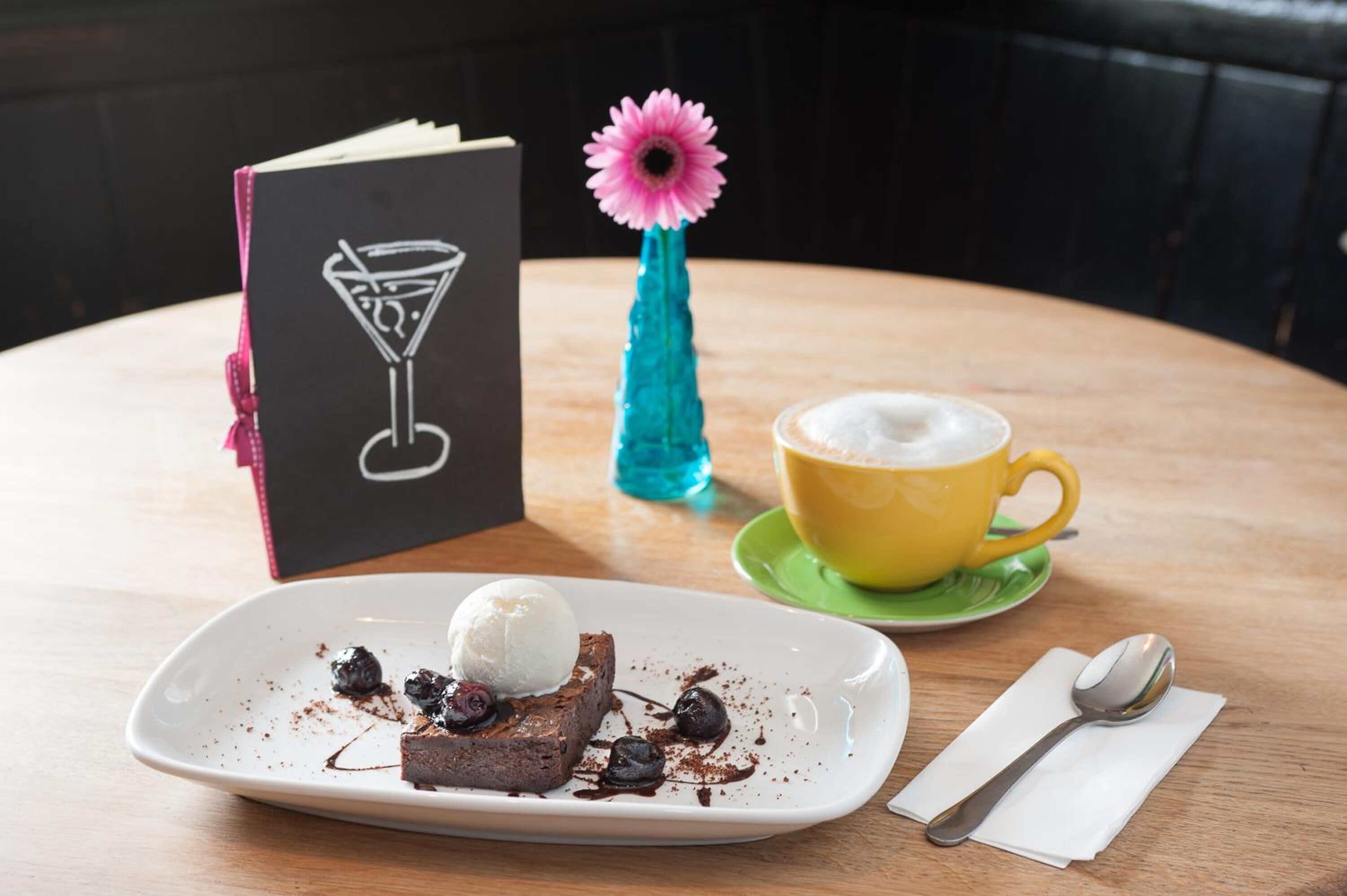 A dessert plate with a scoop of ice cream and a piece of cake, a yellow cup of coffee, a black book with a martini glass drawing, and a blue vase with a pink flower on a wooden table.
