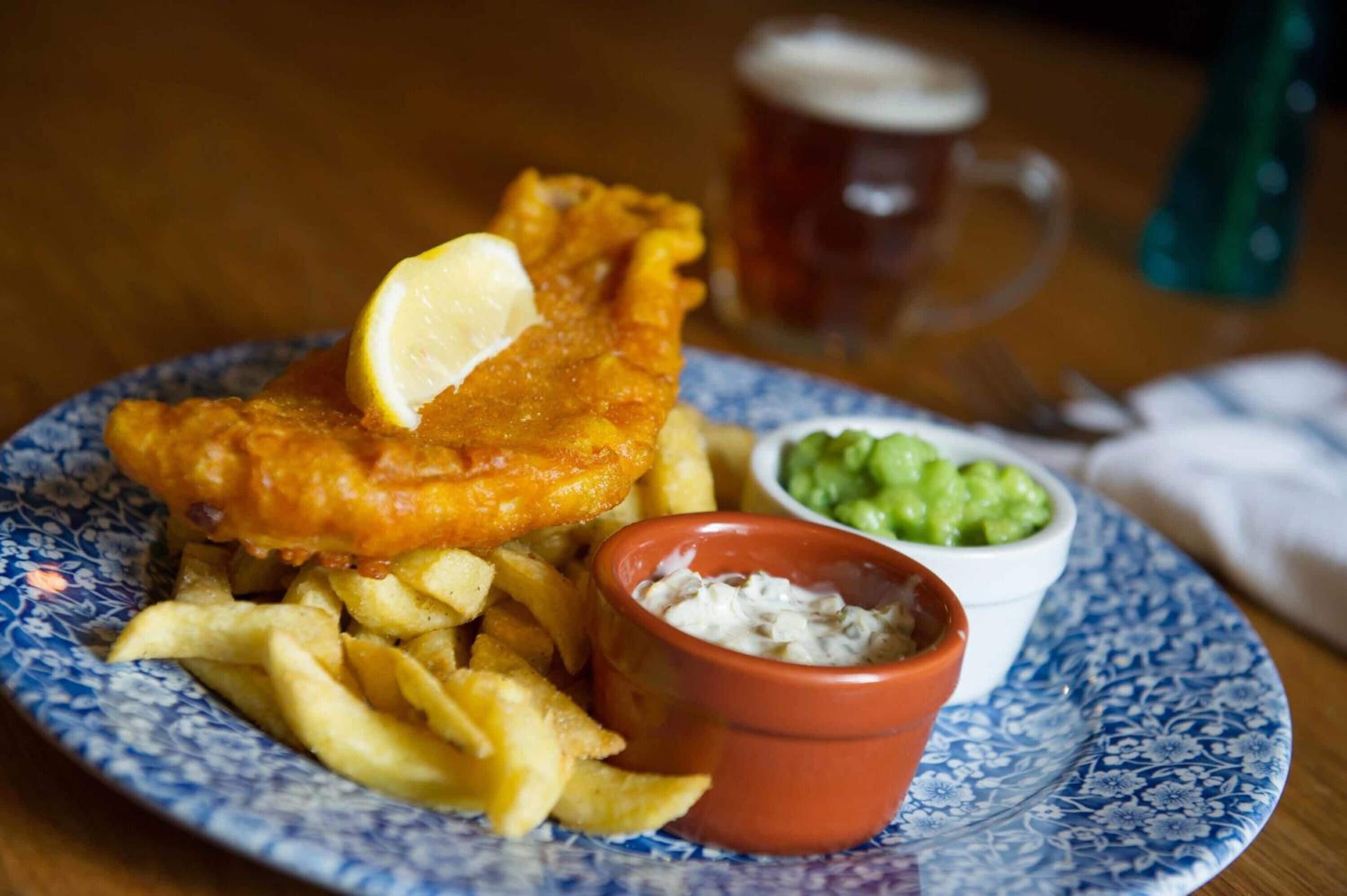 A plate of fish and chips with a lemon wedge, a side of mushy peas, and a small bowl of tartar sauce.