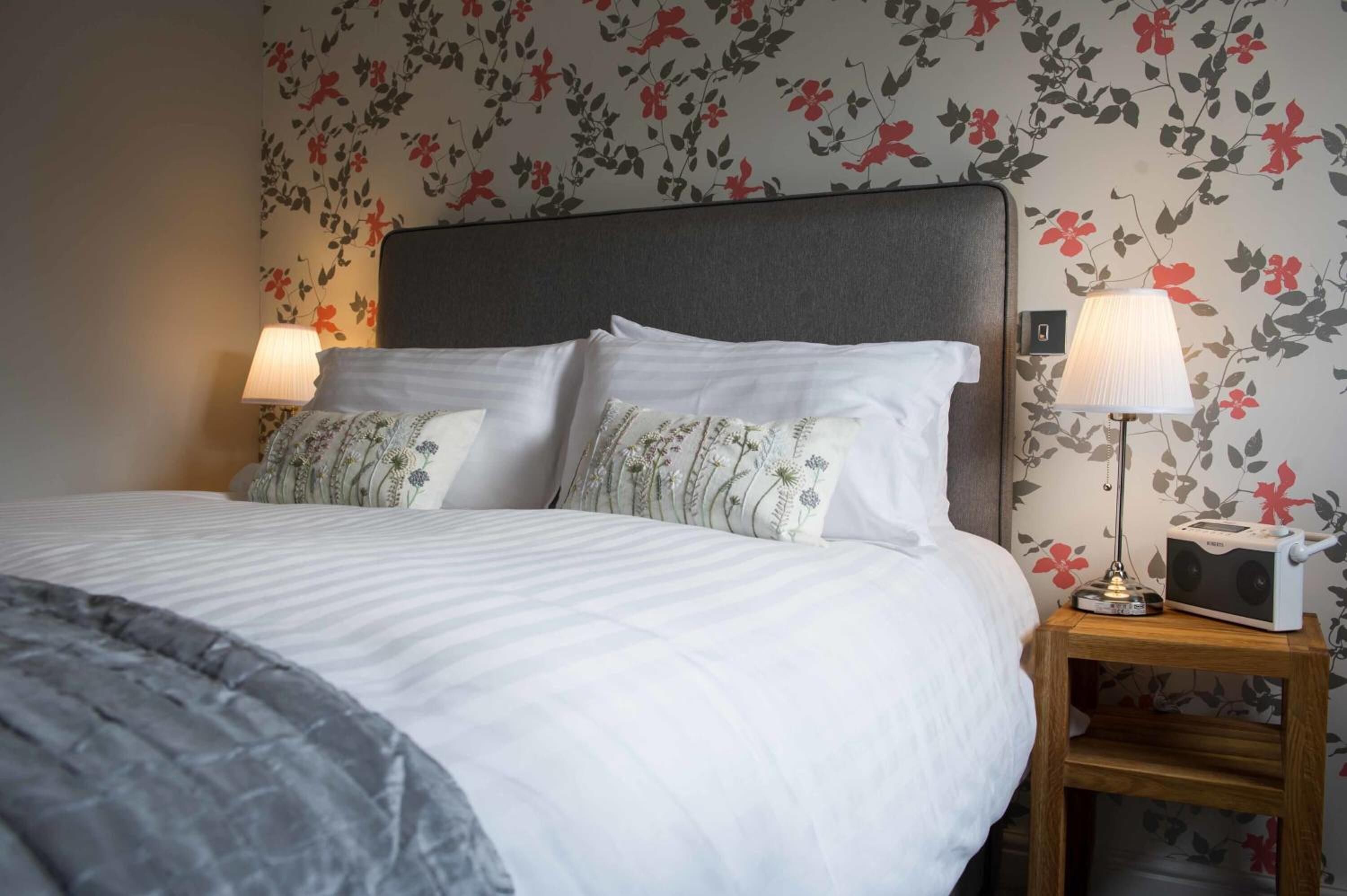 Ein ordentlich bezogenes Bett mit weißen Leinentüchern und Kissen mit Blumenmuster, ein Nachttisch mit Lampe und eine Wand mit floralem Tapetenmuster.