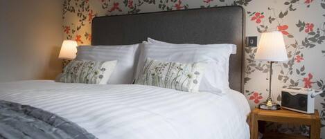 Ein ordentlich bezogenes Bett mit weißen Leinentüchern und Kissen mit Blumenmuster, ein Nachttisch mit Lampe und eine Wand mit floralem Tapetenmuster.