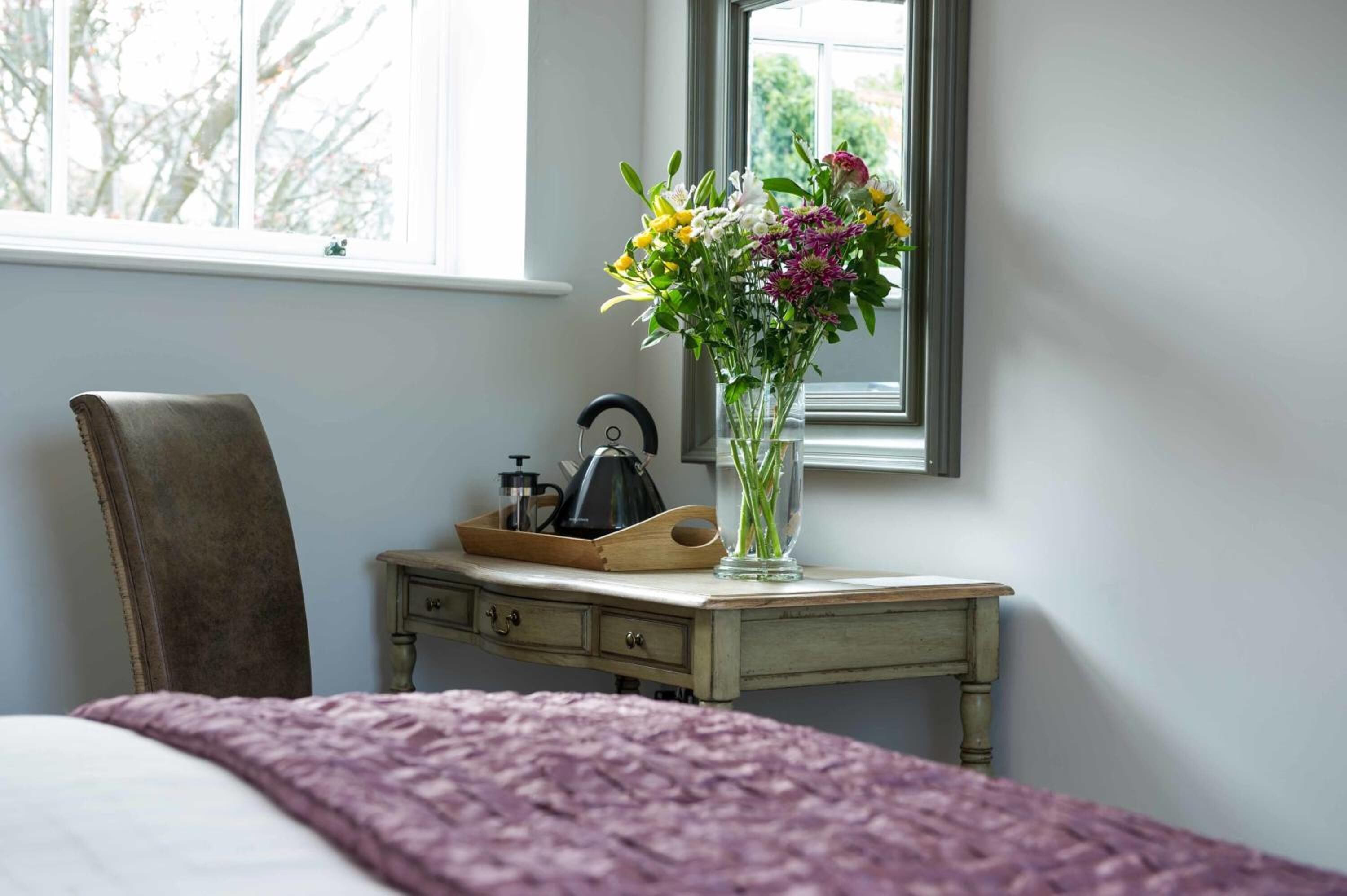 A bedroom with a bed, a chair, a bedside table with a vase of flowers, and a window with a view of trees.