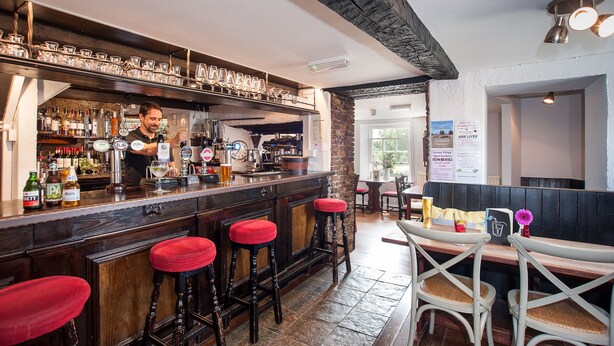 A bar area with stools, a bartender, and a variety of bottles and glasses on the shelves. - Hare & Hounds (Kendal)
