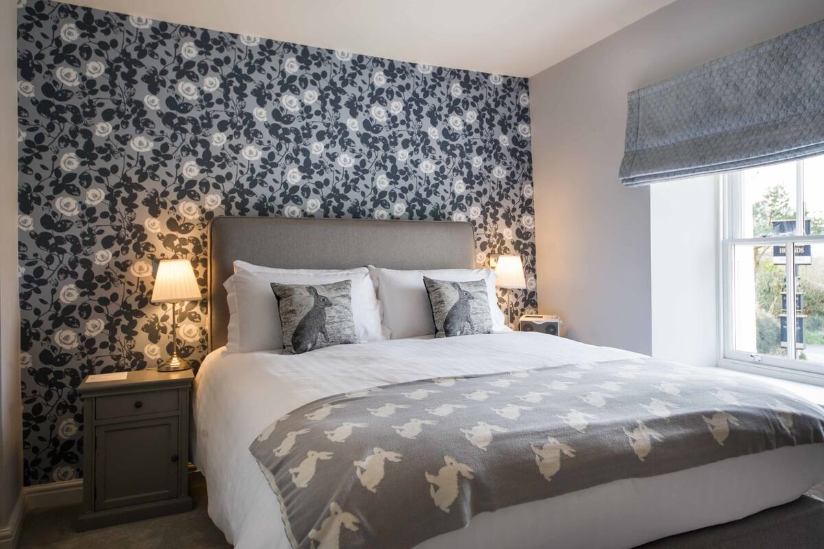 Una cama bien hecha con un cabecero estampado con motivos florales, dos lámparas de noche y una ventana con vistas al exterior.