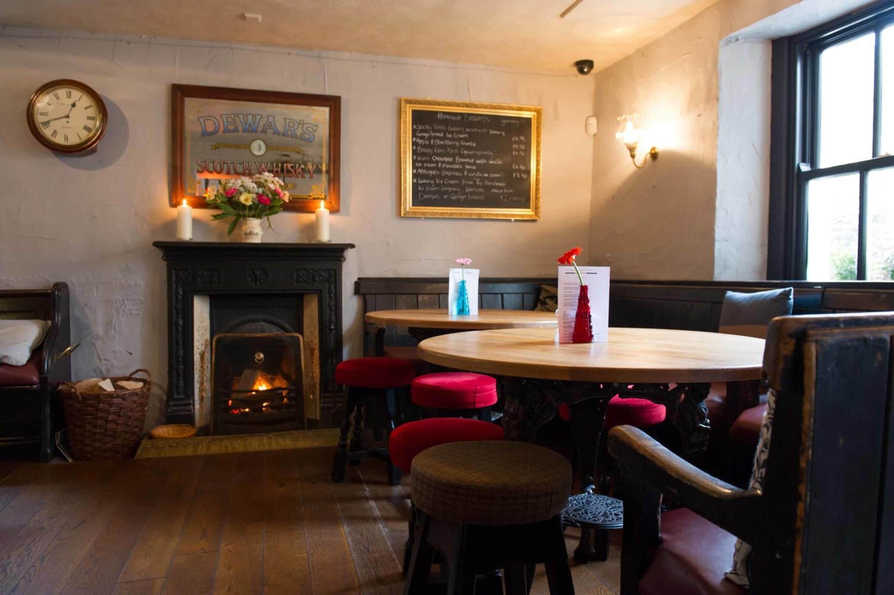 A cozy pub interior with a fireplace, wooden tables, red cushioned stools, and a menu board on the wall.