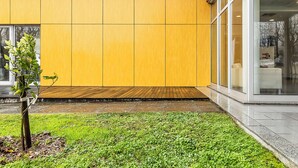 Un edificio moderno con fachada amarilla, una terraza de madera y un área verde con un árbol pequeño.