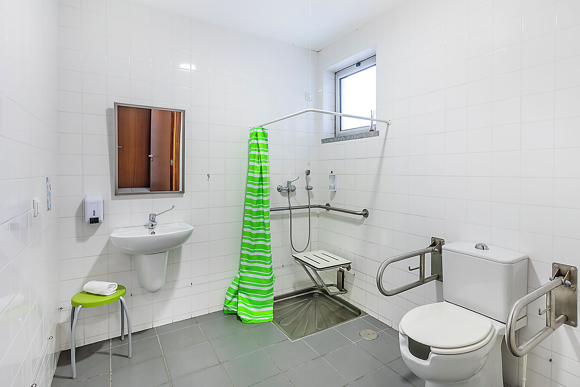 Un baño con un lavamanos blanco, un inodoro, una ducha con cortina verde a rayas y una ventana sobre el inodoro.