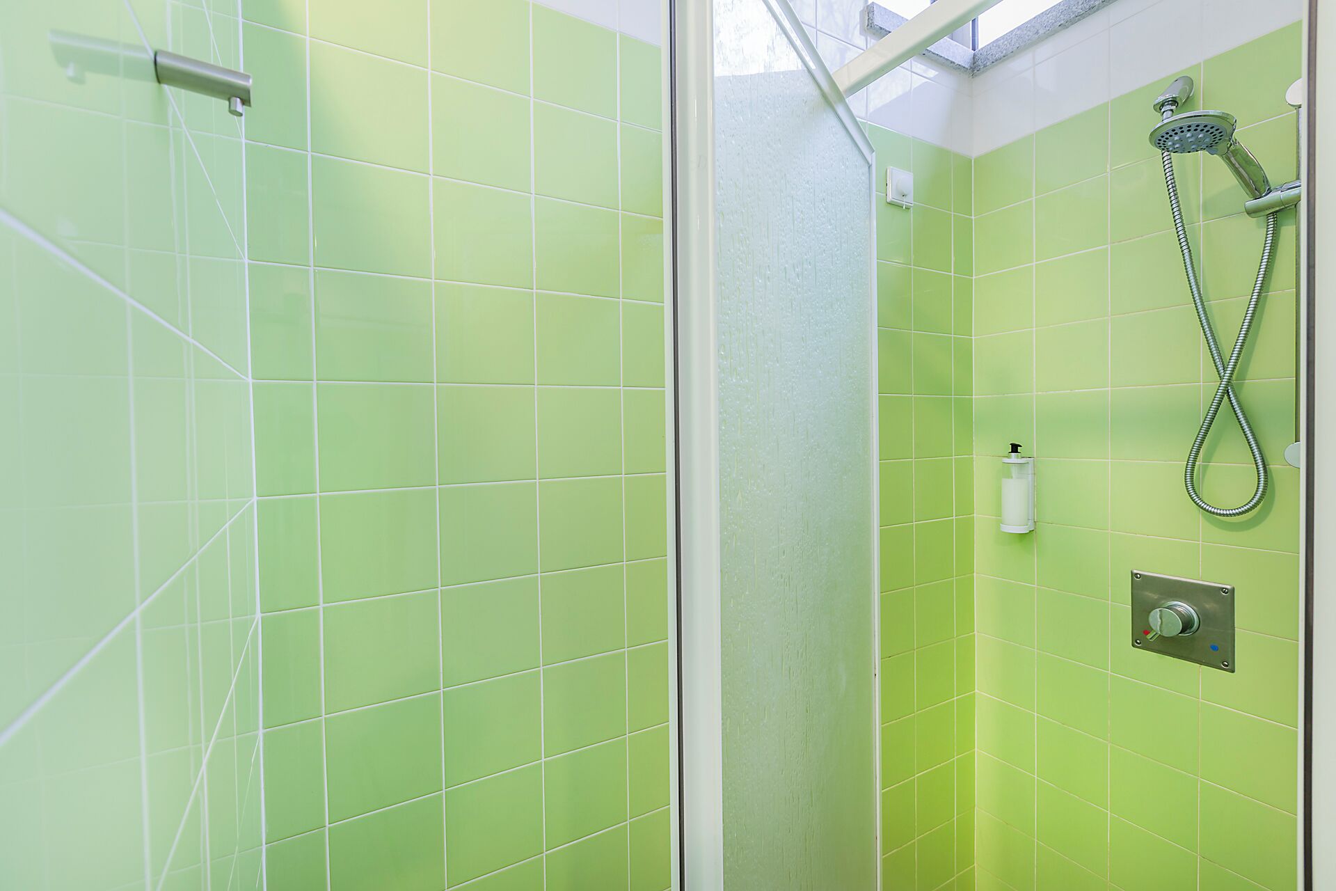 A bathroom with green tiles, a glass shower enclosure, a handheld showerhead, and a wall-mounted faucet.