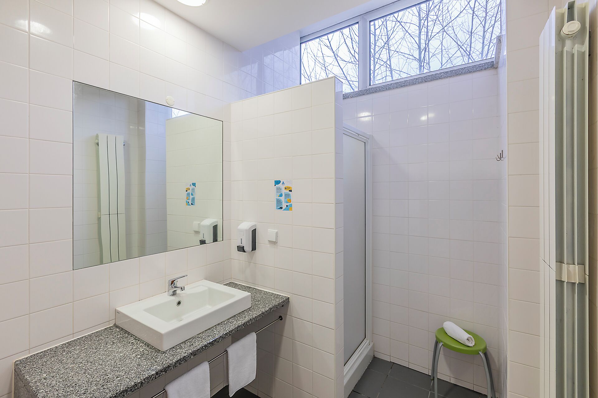 Un baño con un lavamanos blanco, un espejo, una ducha y un taburete verde con una toalla.
