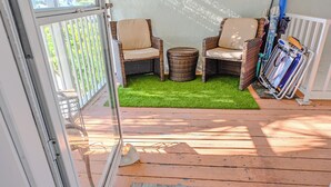 Una terraza de madera con sillas de mimbre, una mesita redonda, un felpudo y un estante metálico con paraguas.