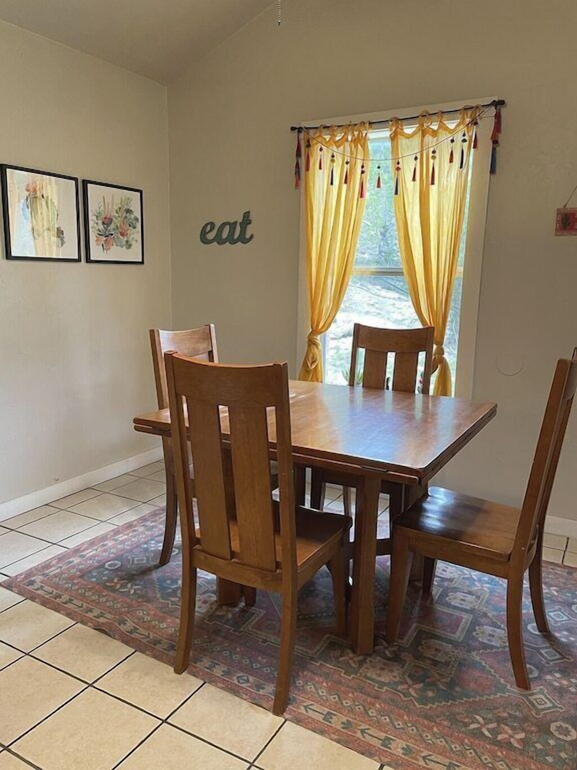 A dining area with a wooden table and four chairs, a patterned rug, and a window with yellow curtains.
