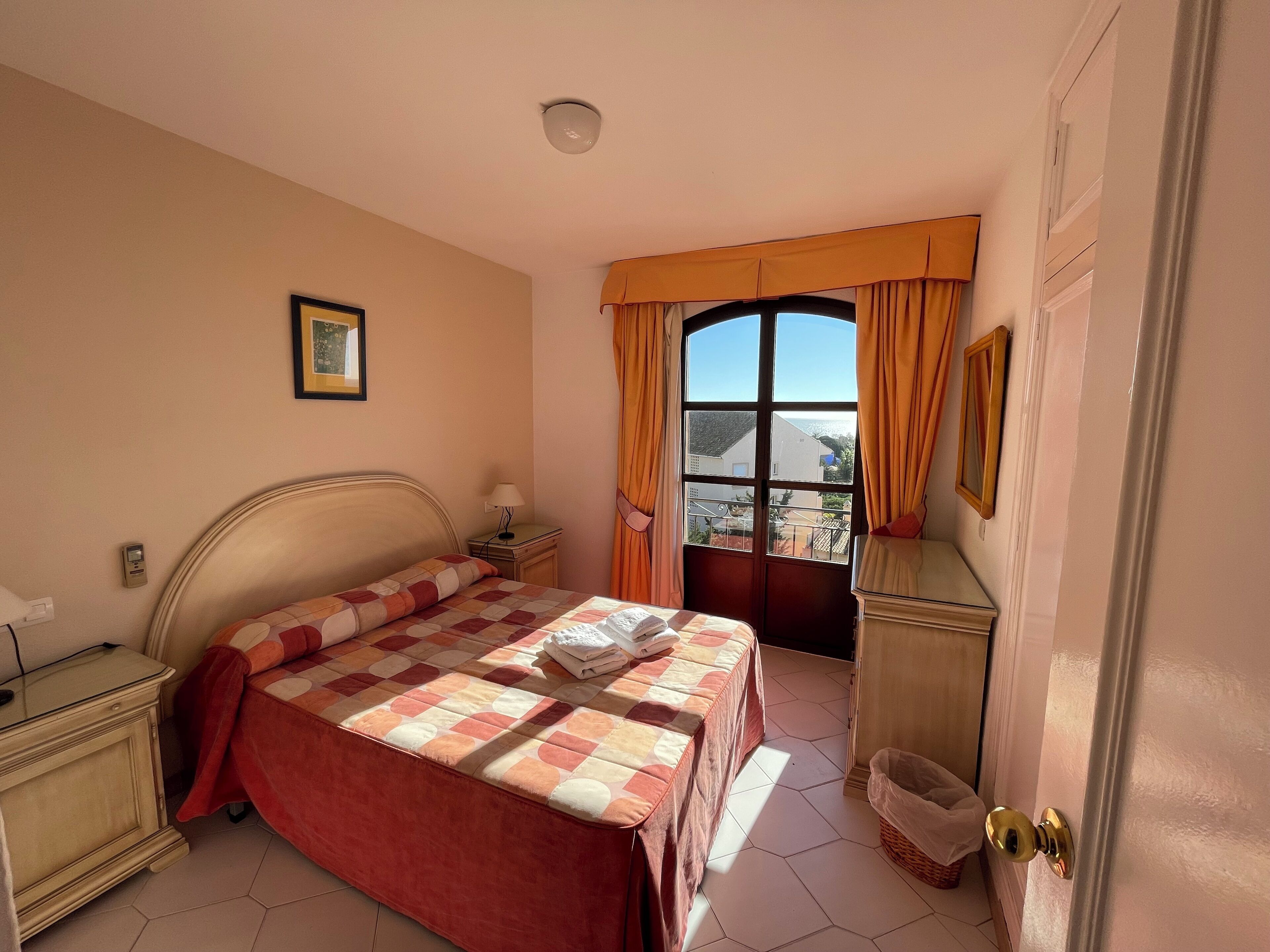 A bedroom with a bed, bedside table, lamp, window with curtains, and a view of buildings outside.