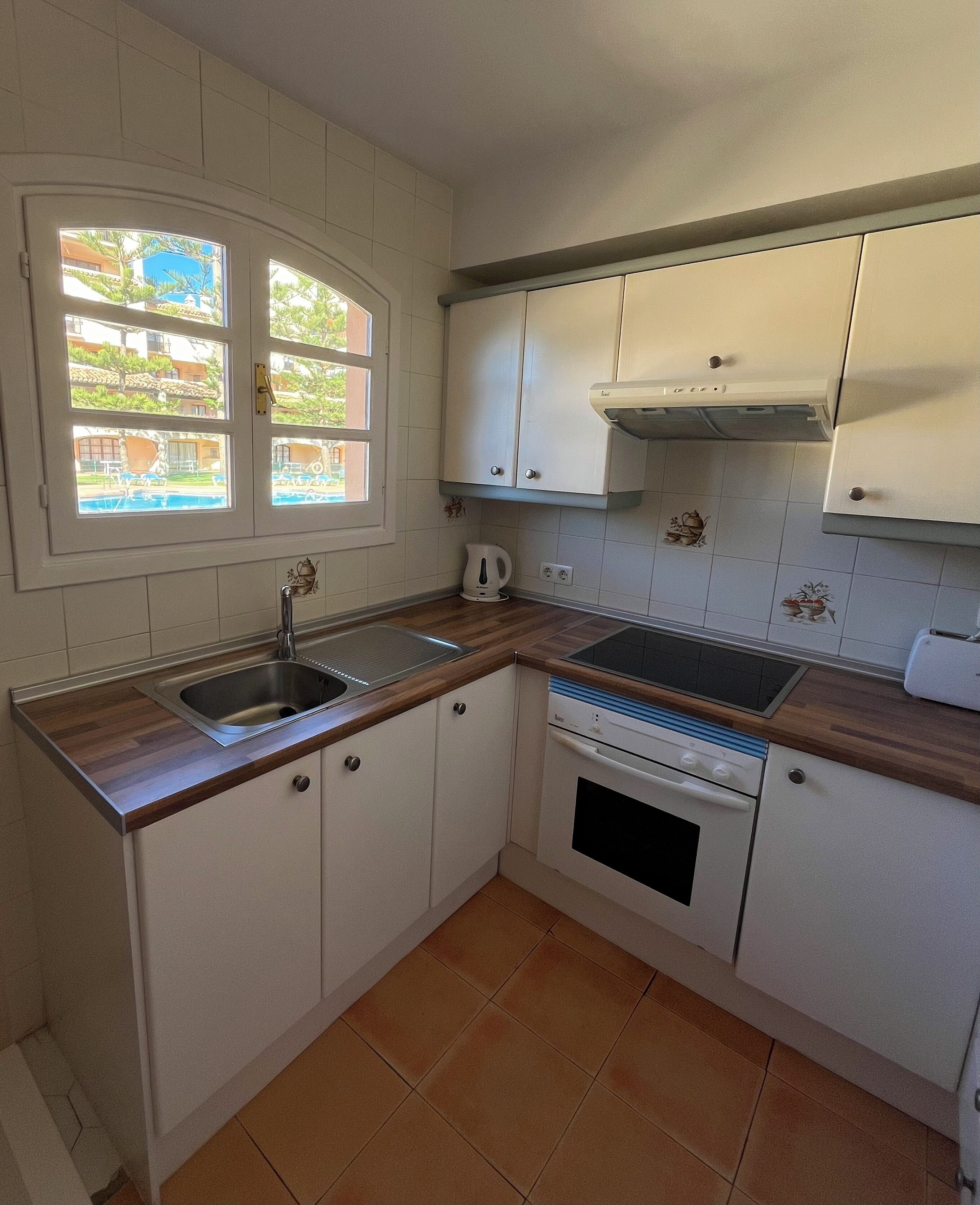 A kitchen with white cabinets, a double sink, a stove, and a window with a view of a pool and buildings.