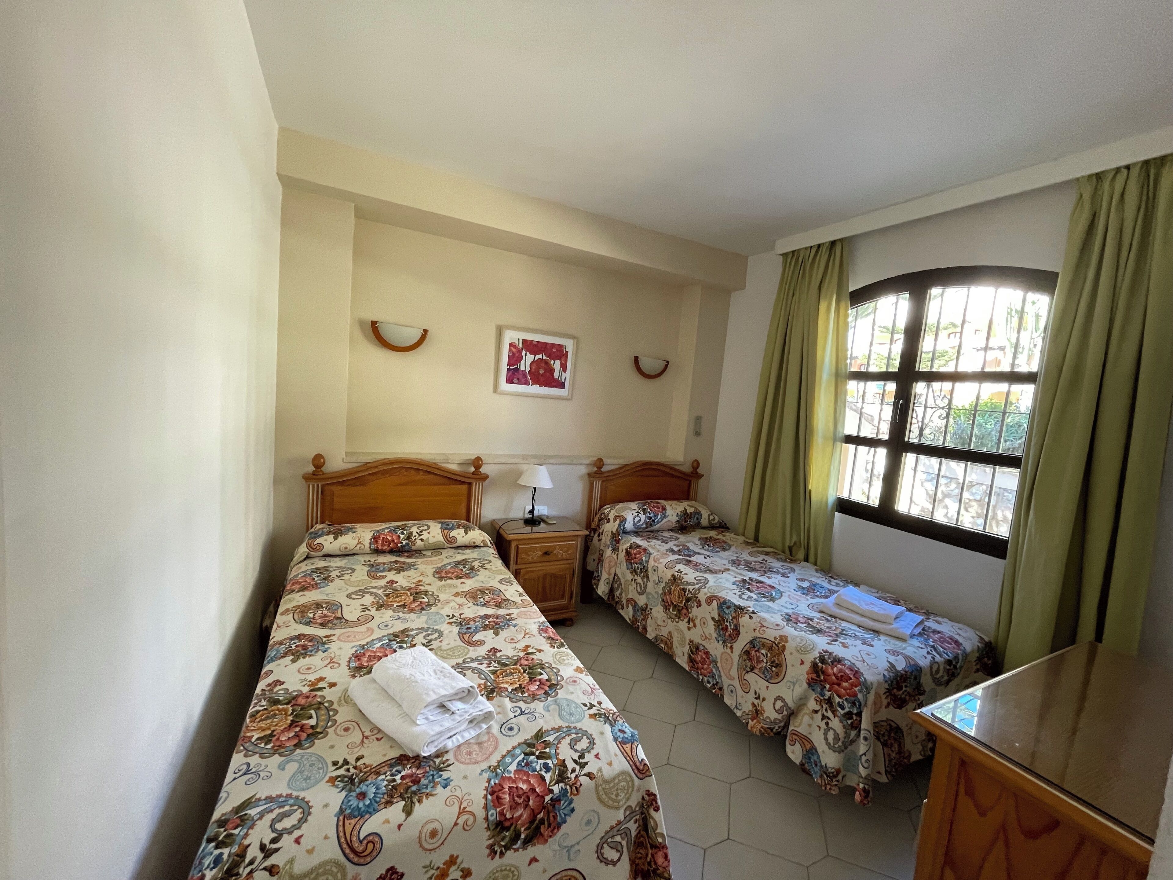 A room with two beds, a bedside table with a lamp, a window with curtains, and a framed picture on the wall.
