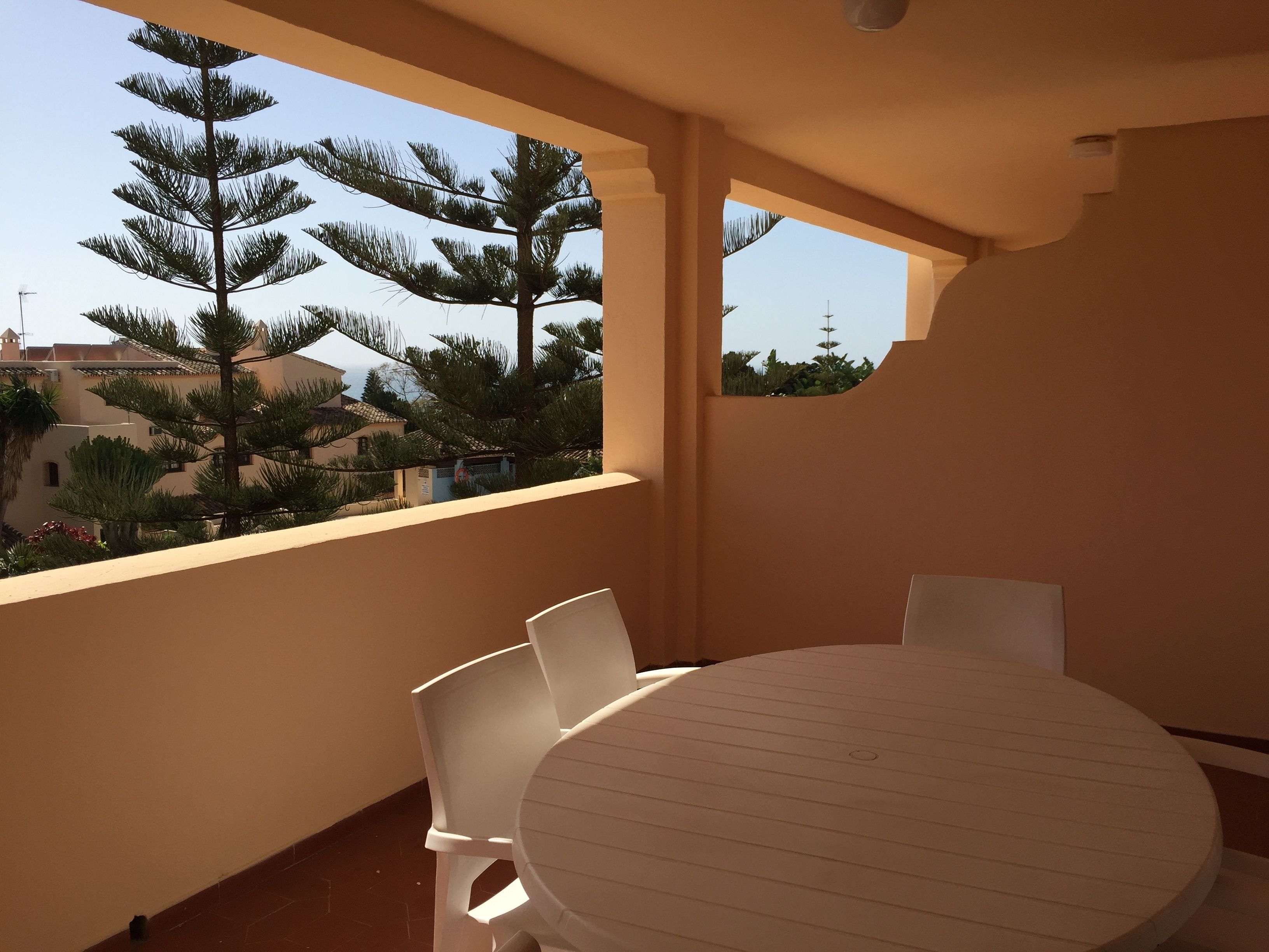 Un balcon avec une table ronde et des chaises, donnant sur un quartier résidentiel bordé d’arbres et de maisons.