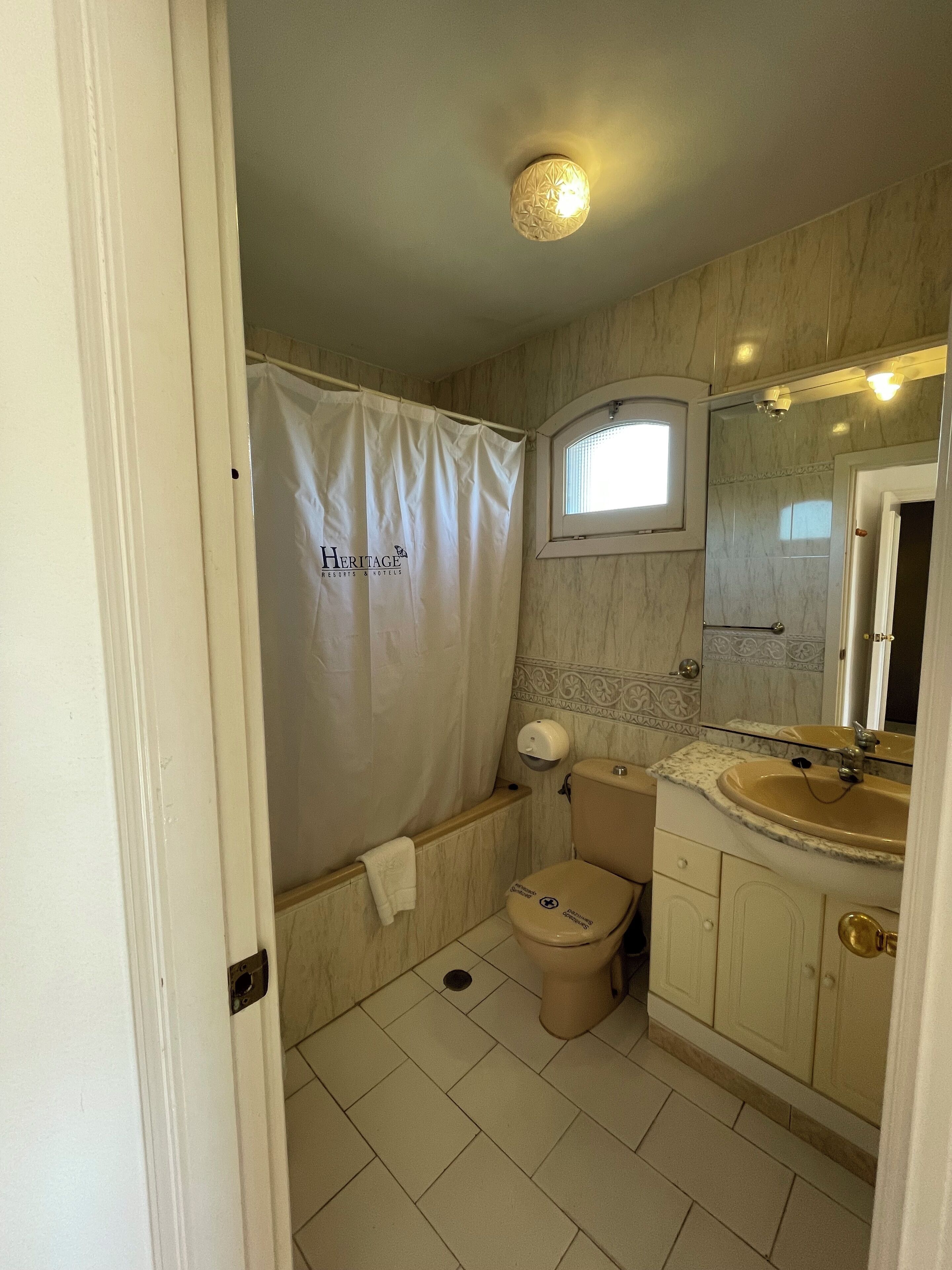 A bathroom with a shower curtain labeled "Heritage," a toilet, a sink with a mirror, and tiled flooring.