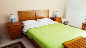 Uma cama bem arrumada com colcha verde, cabeceira de madeira, duas mesas de cabeceira com abajures, uma pequena mesa de madeira e um tapete estampado no chão.