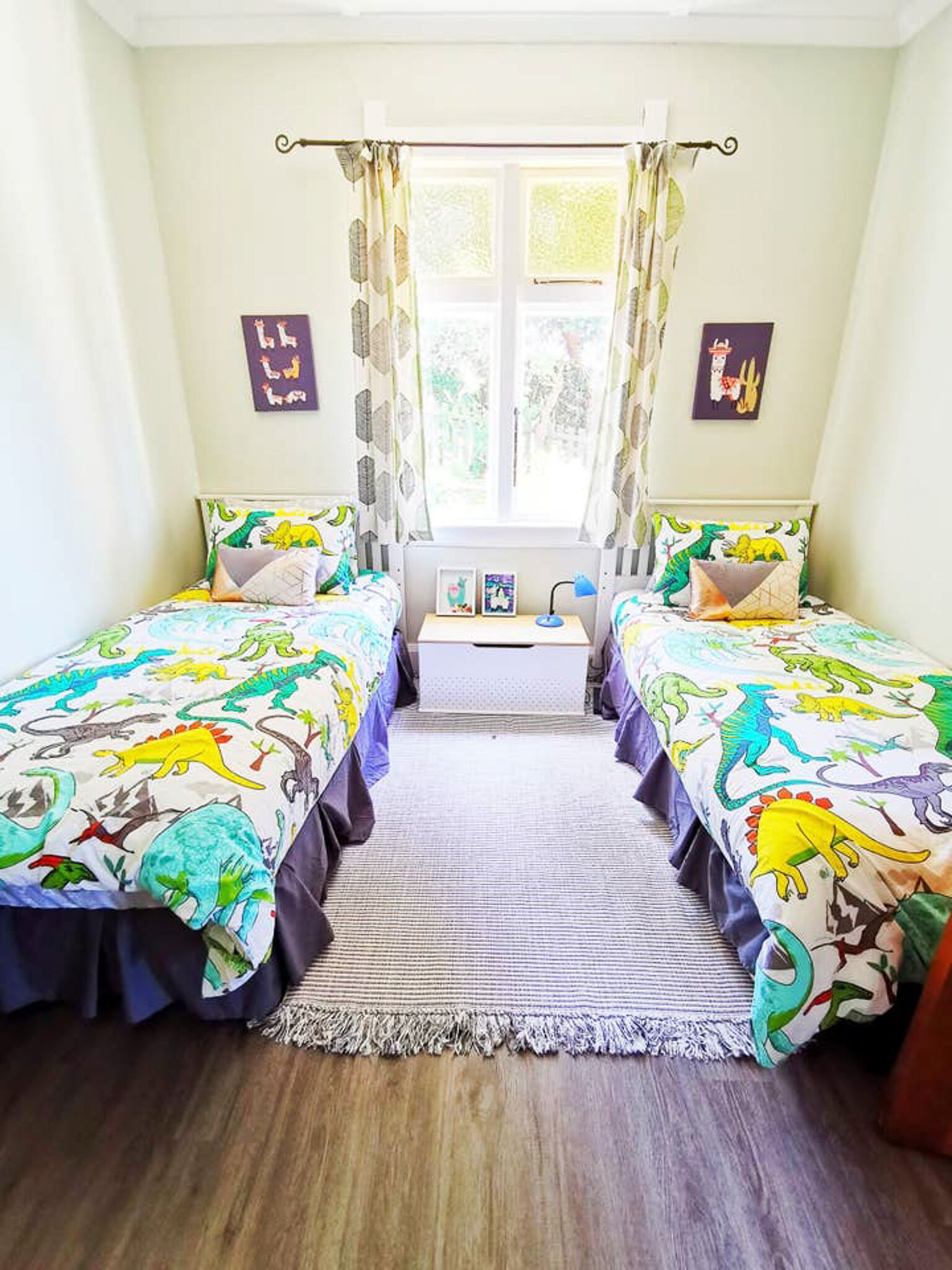 A bedroom with two beds, each with a dinosaur-themed bedspread, a window with curtains, and a small bedside table with a lamp.