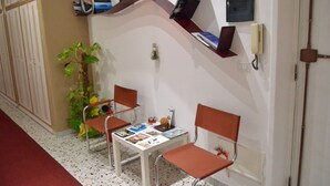 A waiting area with two chairs, a table with magazines, a plant, and a wall-mounted shelf with books and a phone.