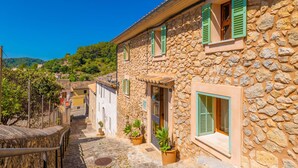 Ein Steinhaus mit grünen Fensterläden, eine enge Gasse und eine Hügelkulisse im Hintergrund.