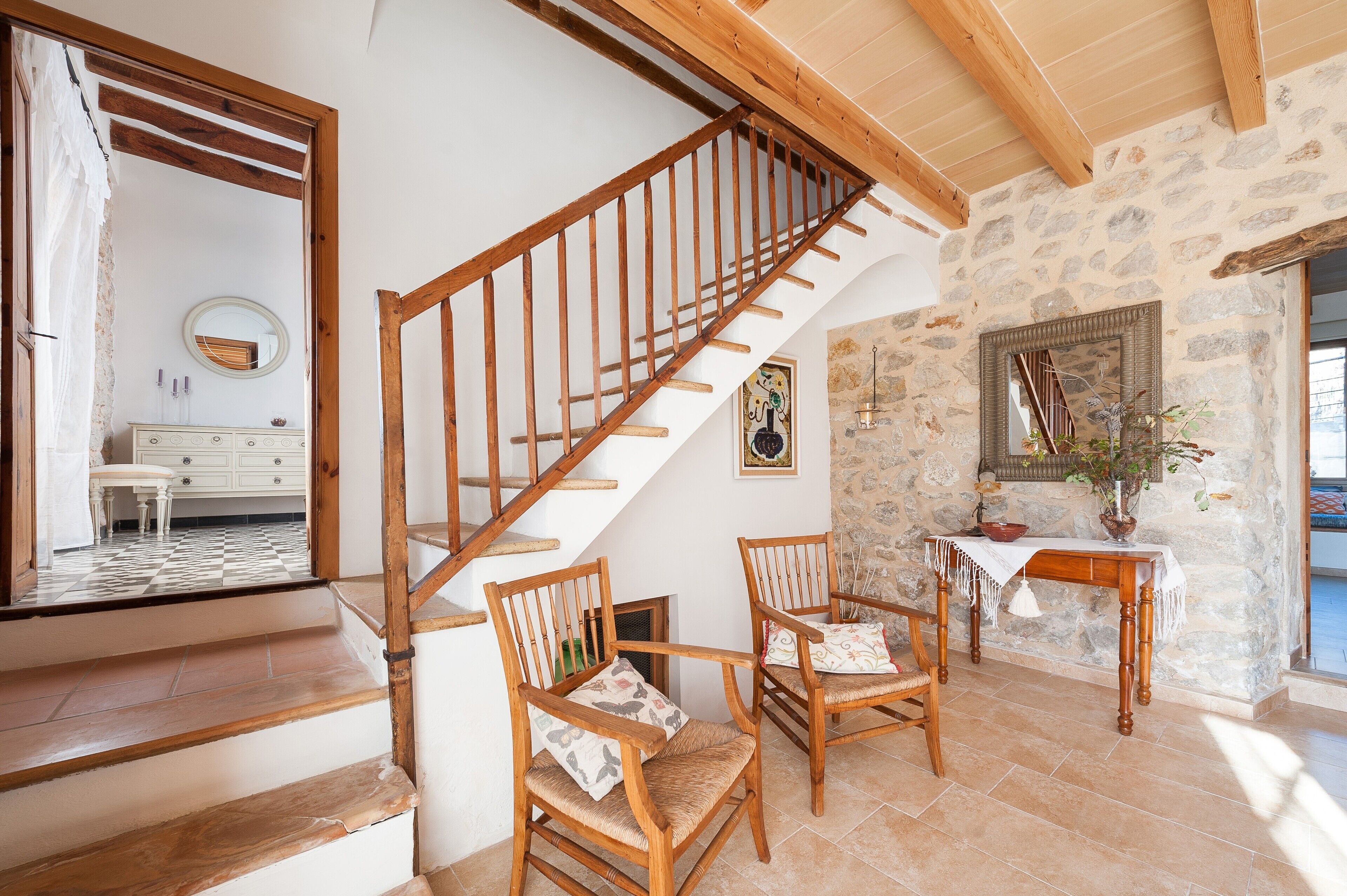 A staircase leading to a room with a stone wall, a wooden table with a vase, and two wooden chairs.