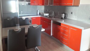 A modern kitchen with red cabinets, a stainless steel countertop, and a dining area with a table and chairs. - Apartments Ivanka (Okrug)