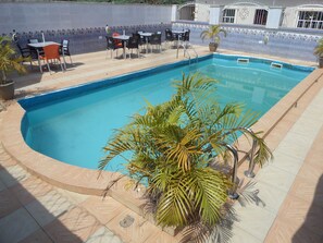 Una piscina rectangular con borde de azulejos, rodeada por un patio con baldosas y mobiliario exterior.