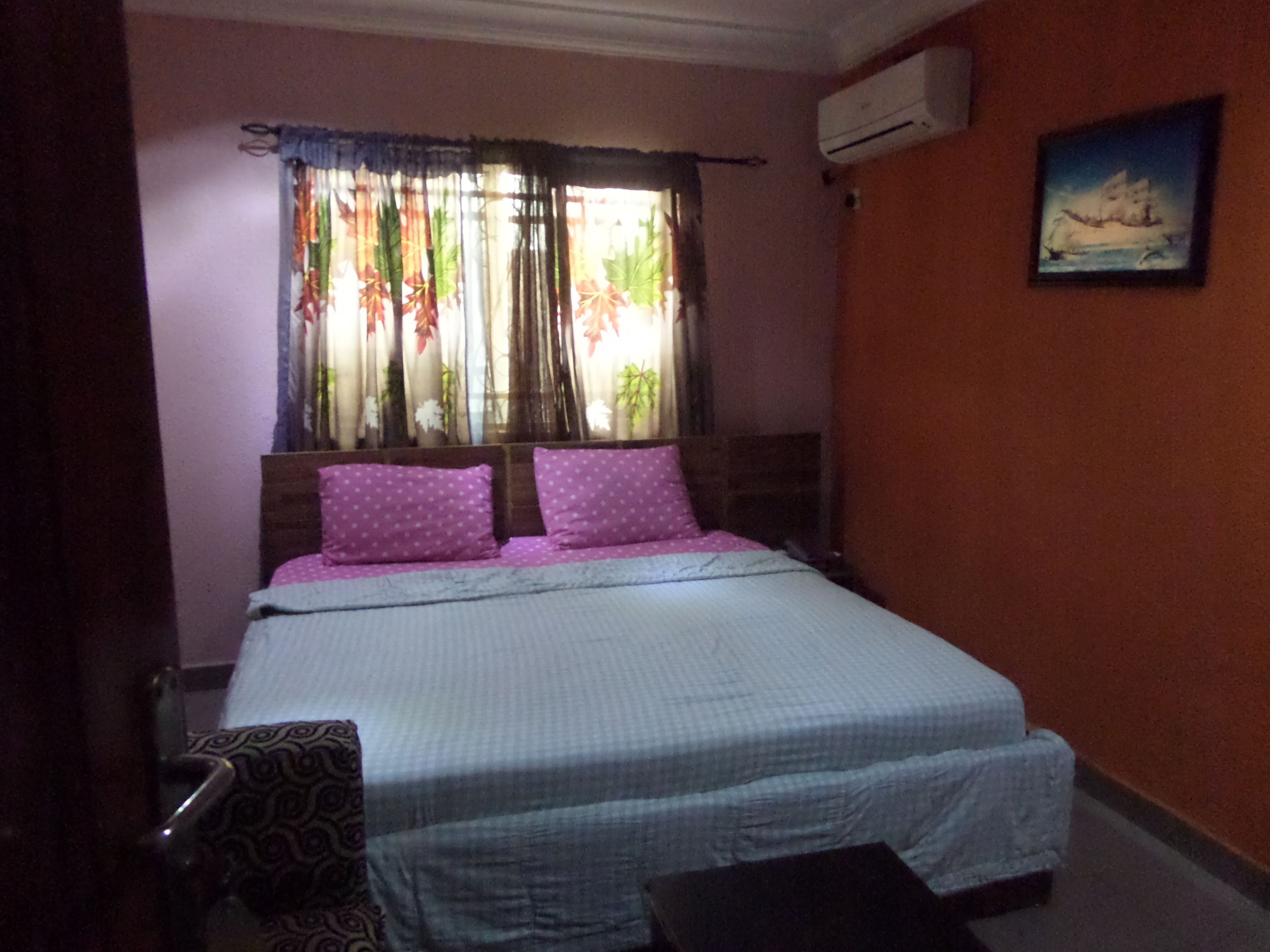 A bedroom with a bed, pillows, a window with curtains, an air conditioner, and a painting on the wall.