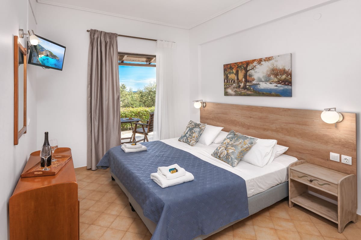 A hotel room with a bed, a wooden headboard, a TV mounted on the wall, a painting, and a window with curtains.