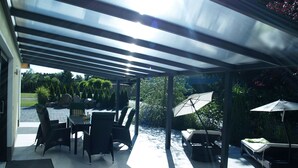 Ein überdachter Patio-Bereich mit Gartenmöbeln, darunter ein Tisch, Stühle und eine Liege mit Sonnenschirm.