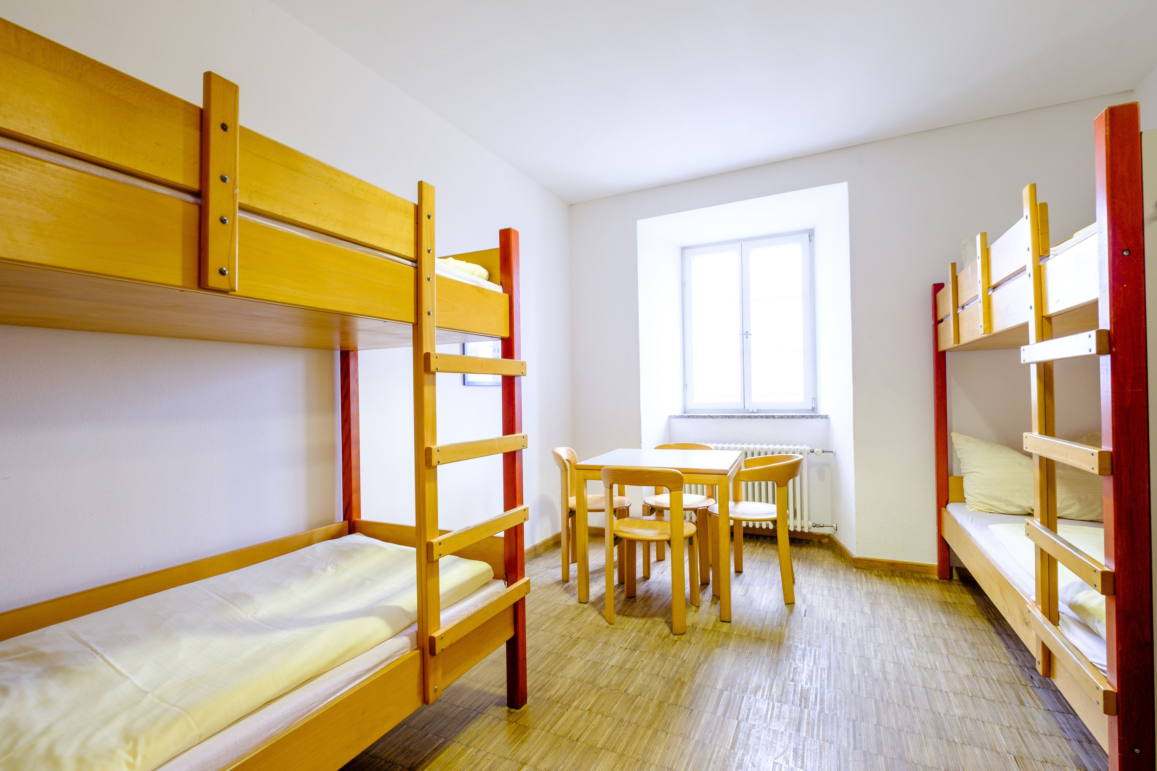 A dormitory room with bunk beds, a wooden table, chairs, and a window.