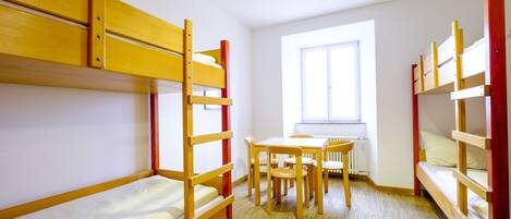 A dormitory room with bunk beds, a wooden table, chairs, and a window.