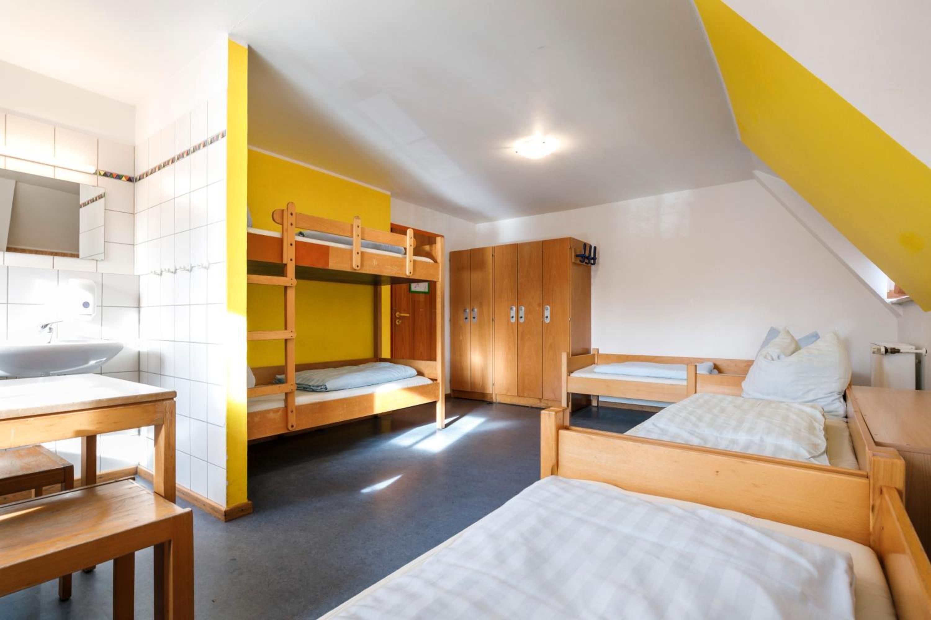 A dormitory room with bunk beds, a wooden table, a sink, and a wardrobe.