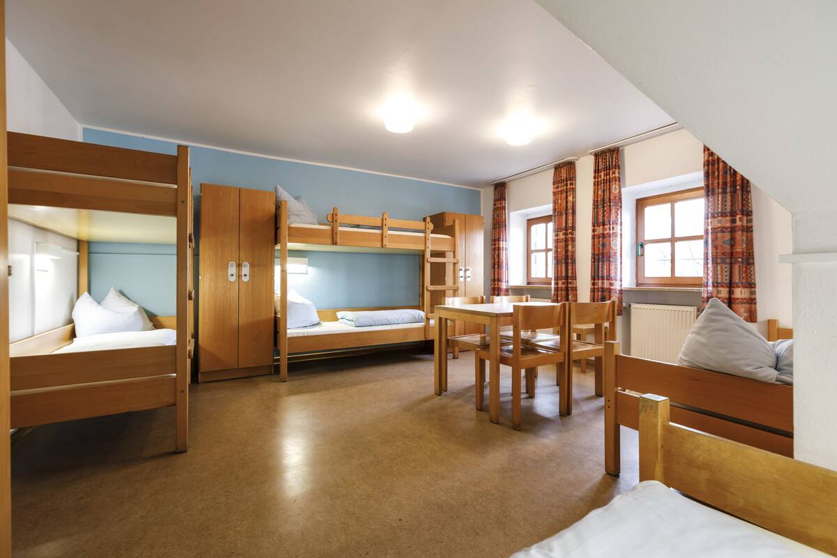 Ein Schlafsaalzimmer mit Etagenbetten, einem Schreibtisch, Stühlen und einem Fenster mit Vorhängen.