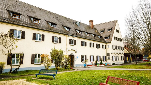 Ein Gebäude mit mehreren Fenstern, einem geneigten Dach und einer gepflasterten Fläche mit Bänken.