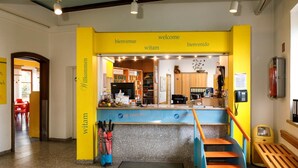 Ein Empfangsbereich mit gelb-blauer Farbgestaltung, mit einer Rezeption, einem Telefon und einer kleinen Vitrine. Eine Treppe führt zur Rezeption, und rechts ist eine Bank zu sehen.