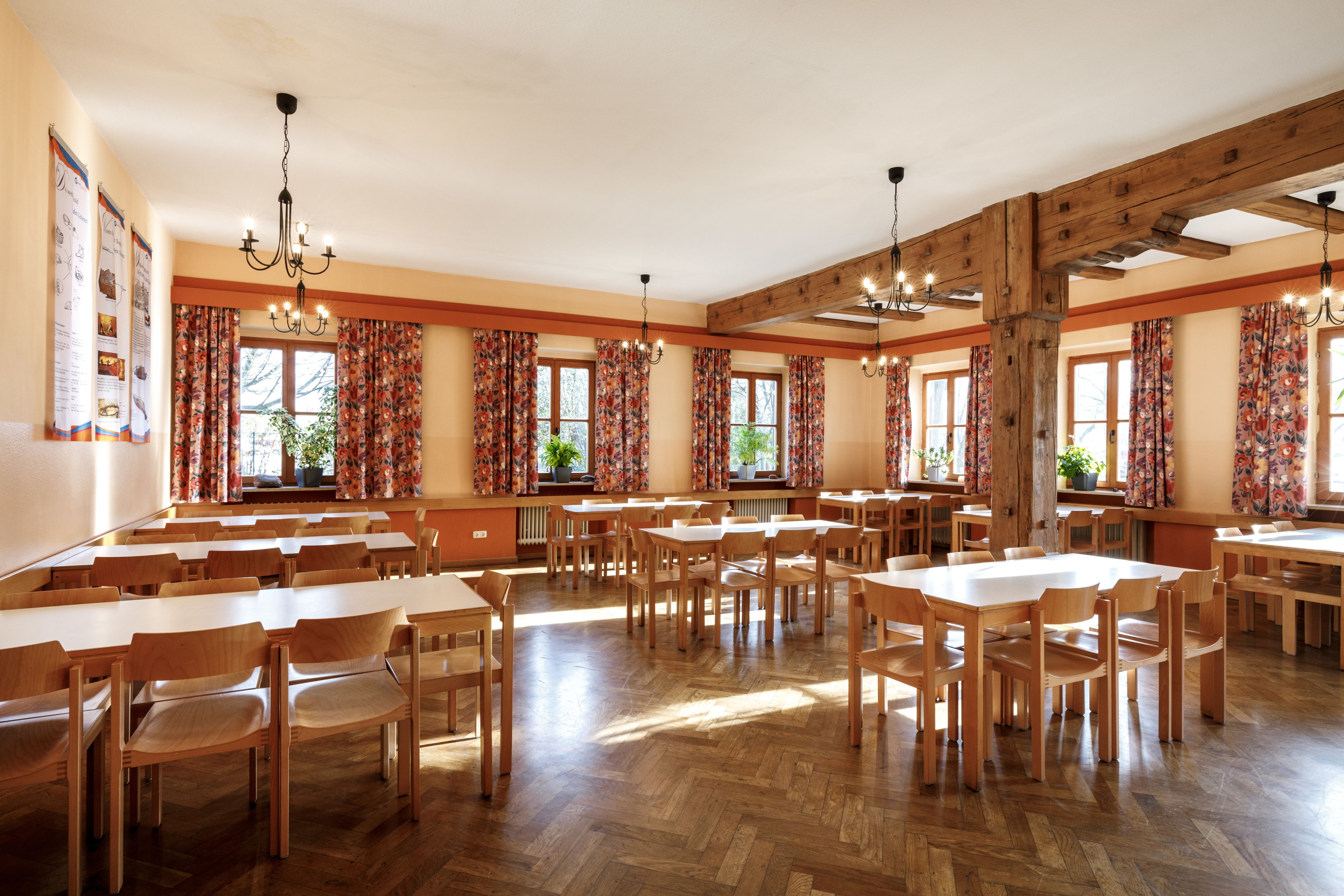 A spacious dining hall with wooden tables and chairs, large windows with floral curtains, and exposed wooden beams on the ceiling.
