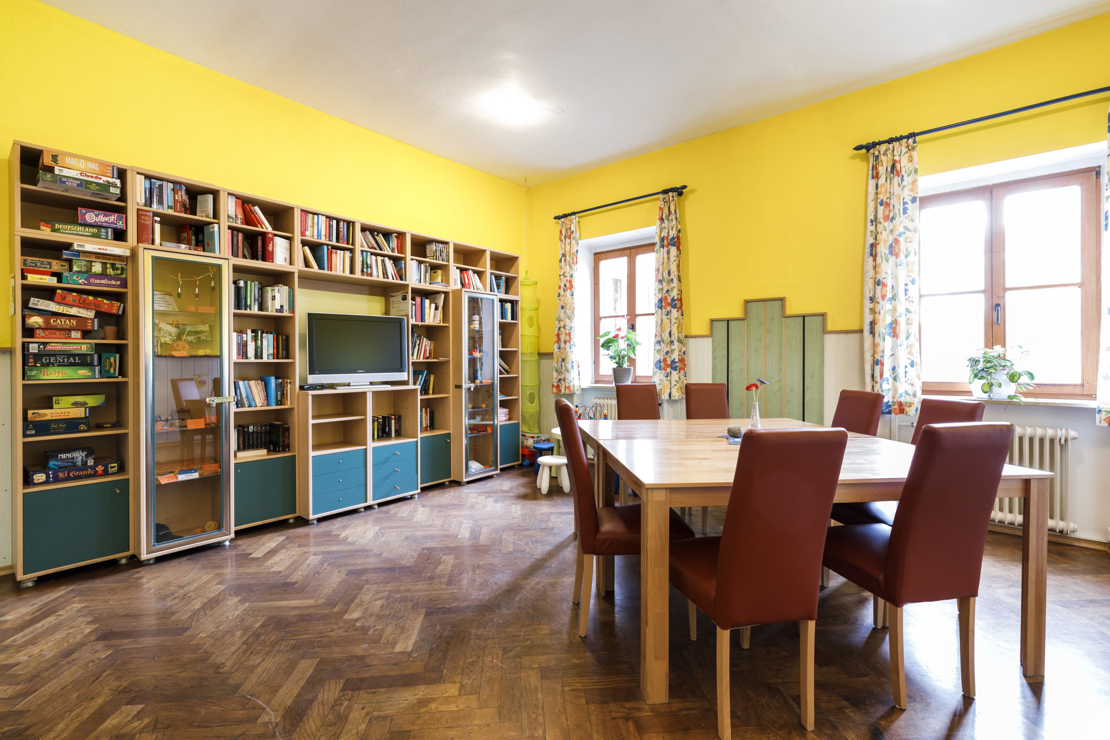A room with a wooden herringbone floor, a dining table with six chairs, and a bookshelf filled with various books and games.