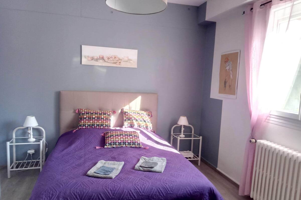 A bedroom with a bed, two bedside tables, a radiator, and a window with curtains.