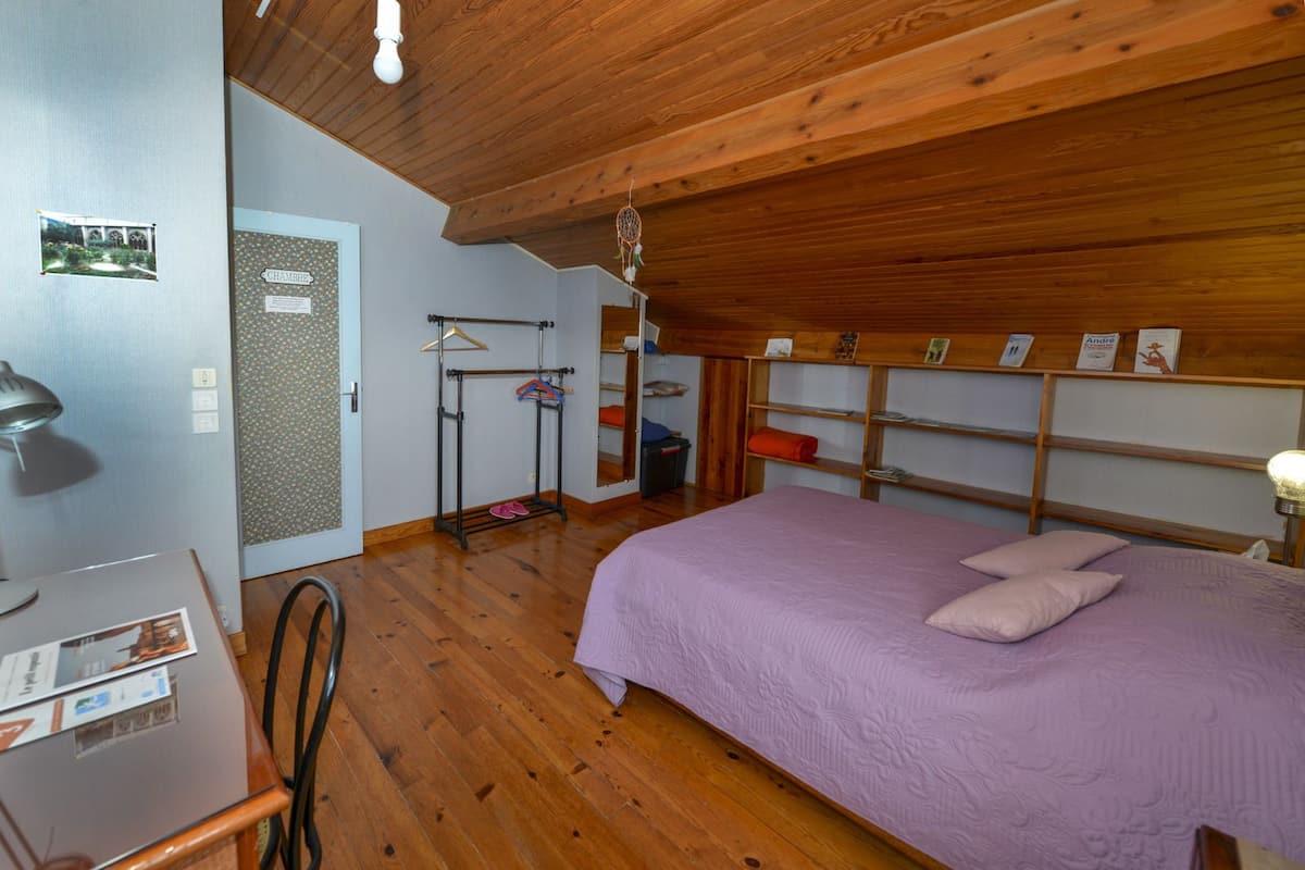 A bedroom with a wooden ceiling, a bed with a purple bedspread, a desk with a lamp, and a coat rack with hangers.